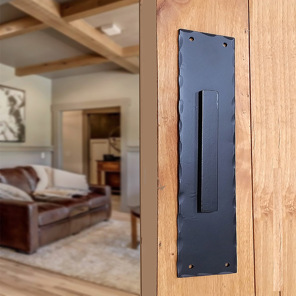 A photo of a large, black flushed finger pull handle with hammered edges installed on a wooden door.